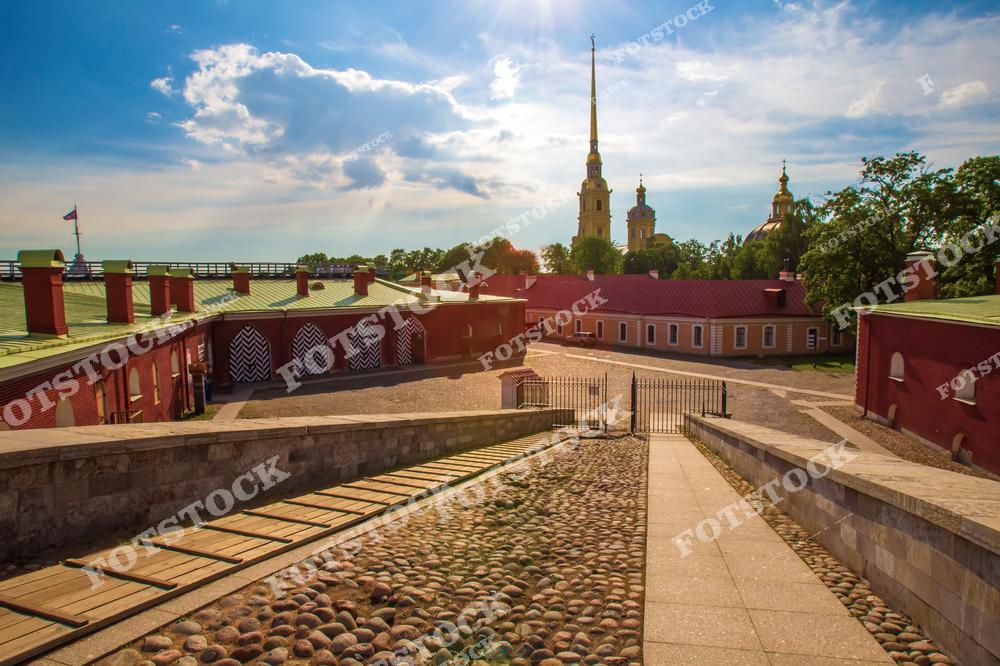 На изображении изображена Петропавловская крепость в Санкт-Петербурге. Виден брусчатый двор перед красными зданиями крепости, украшенными арками. Вдалеке виднеется золотой шпиль Петропавловского собора под ярким голубым небом с облаками.